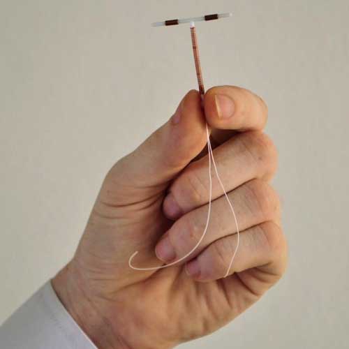 A hand holding an intrauterine device (IUD) with two strings, showcasing the contraceptive tool against a plain background.