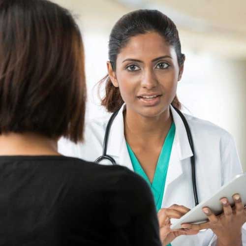 A healthcare professional in a white coat holds a tablet, speaking with a patient in a black top during a consultation.