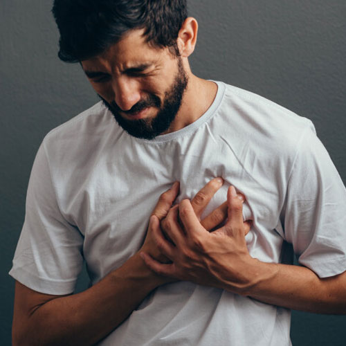 A person in a white t-shirt clutches their chest with both hands, appearing concerned or in discomfort against a gray backdrop.