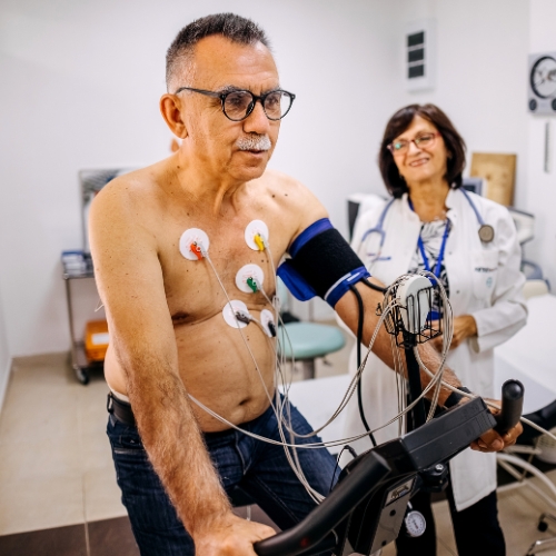 A man on a stationary bike, connected to medical equipment for monitoring, while a healthcare professional observes.