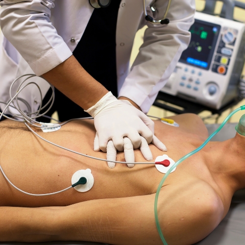 A healthcare professional performs CPR on an unresponsive patient, using a defibrillator with monitoring equipment in the background.