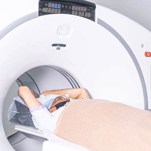 A patient lies on a bed entering a medical imaging machine, likely a CT or MRI scanner, with a blanket covering their body and arms raised above their head.