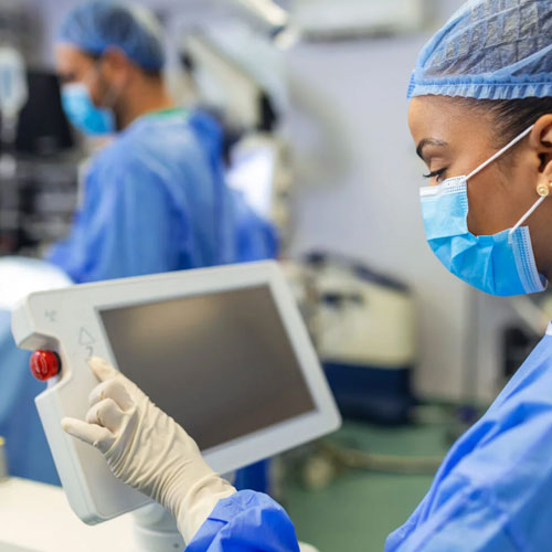 A healthcare professional in scrubs and gloves operates a medical monitor in a surgical setting, focused on the screen's display.