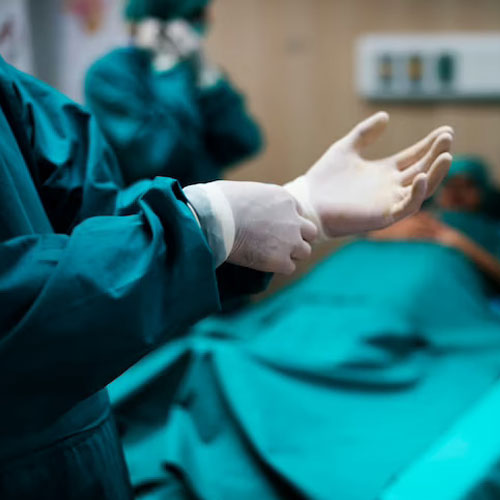 A surgeon in scrubs pulls on a sterile glove in a hospital operating room, with a patient partially visible under a green drape.