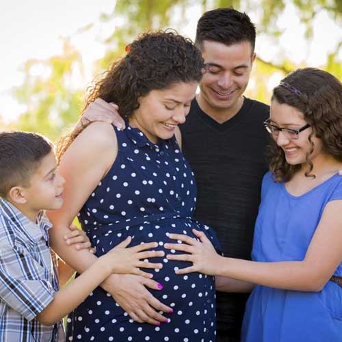 A family of four surrounds a pregnant woman, joyfully touching her baby bump in a sunlit outdoor setting.