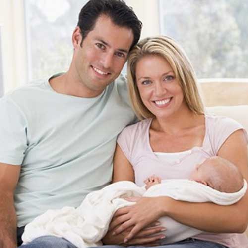 A couple sits close together on a couch, lovingly holding their swaddled baby in a cozy, sunlit living room.