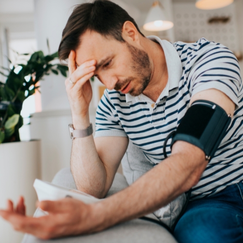 A man in a striped shirt measures his blood pressure using a cuff while sitting on a sofa, looking concerned, with a tablet beside him.
