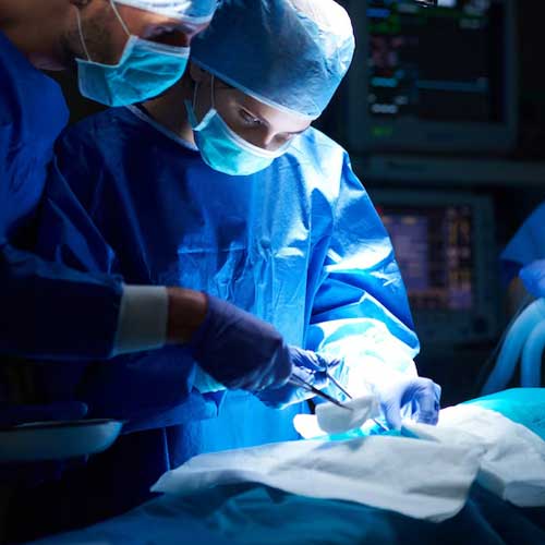 Two medical professionals in blue scrubs and masks perform surgery, focused on a patient under bright surgical lights.