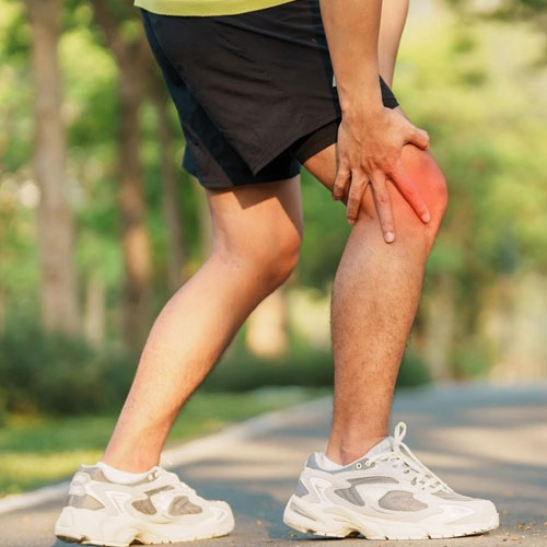 A man in athletic wear holds his knee in pain while standing on a pathway surrounded by greenery.