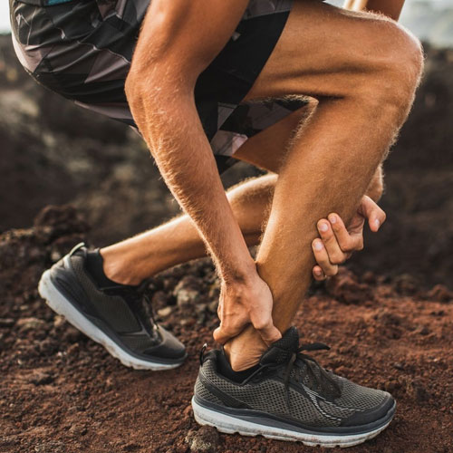 A man in black athletic shoes kneels on rocky ground, gripping his ankle in pain, indicating a possible injury.