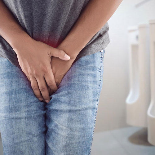 A person in gray shirt and blue jeans holds their groin, indicating discomfort, with restroom stalls in the background.